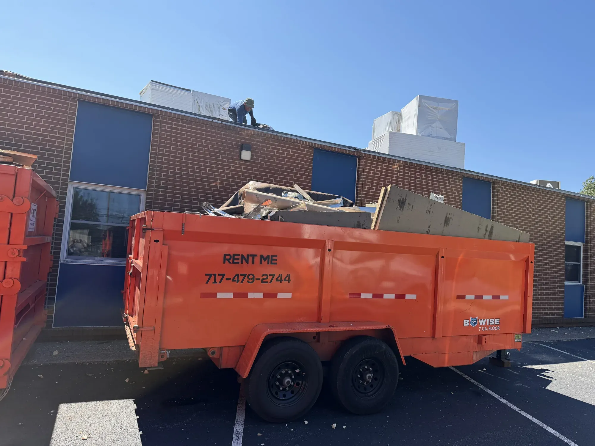 Orange Kingdom Haulers RENT ME trailer dumpster loaded with roofing debris beside a commercial building