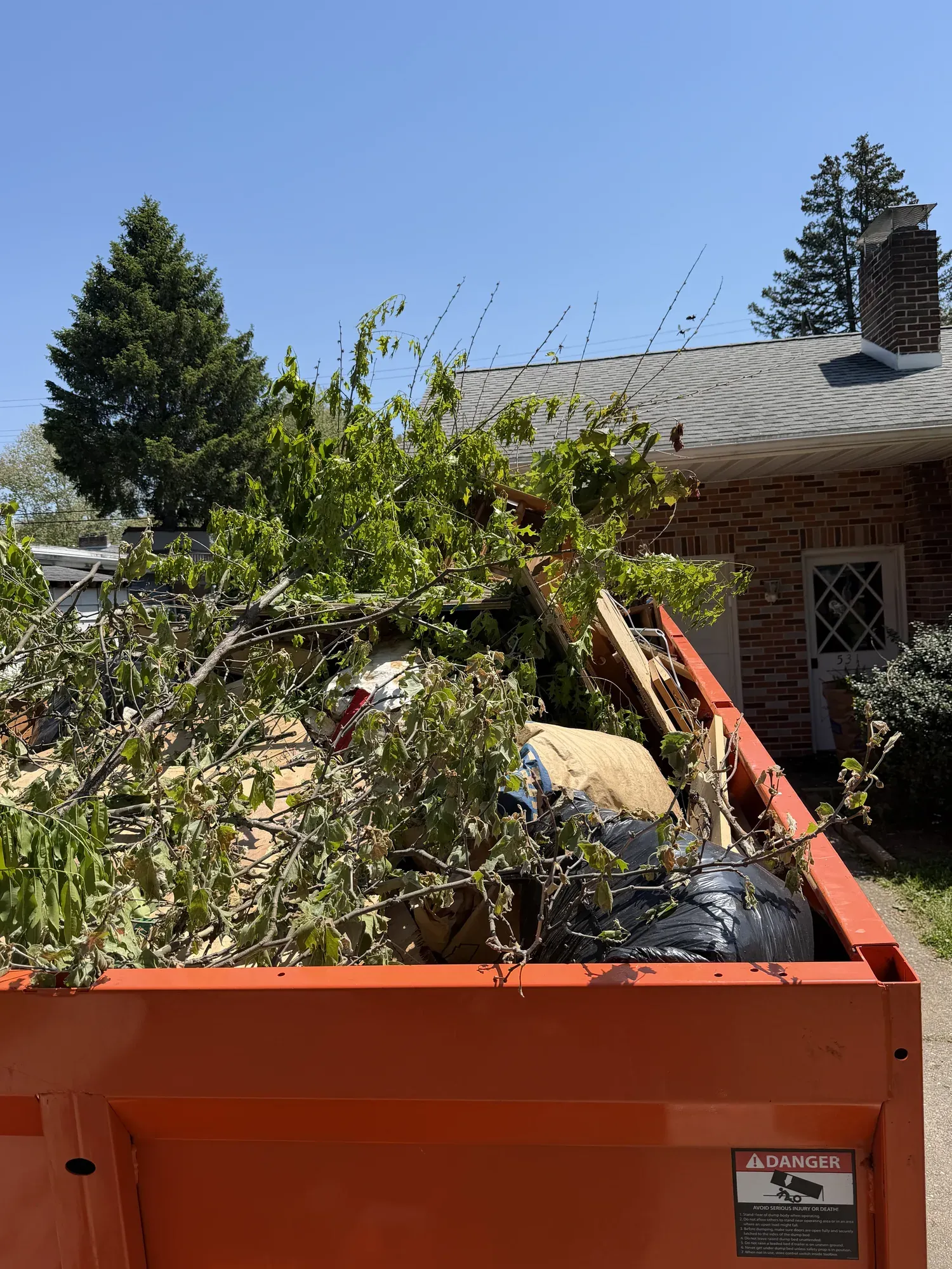 Orange Kingdom Haulers trailer piled with branches, brush and bagged yard waste beside a house